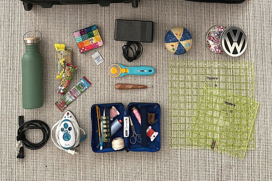 An overhead look at a collection of supplies to bring on a sew day or quilt retreat, including a needle book, travel iron, pins, rotary cutter, and more.