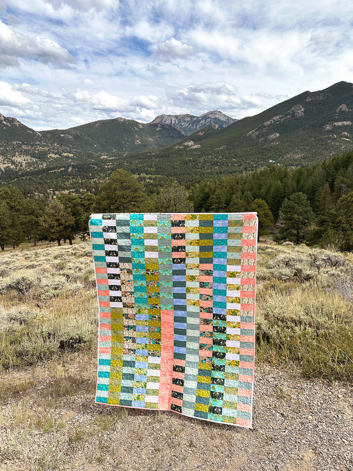 The Due Pinoli Baseline Quilt made with precut jelly roll strips is shown in front of the Rocky Mountains outside Rocky Mountain National Park in Colorado